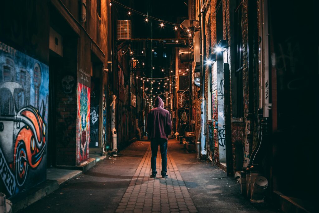 A solitary figure stands in a graffiti-lined alley, illuminated by string lights, at night.