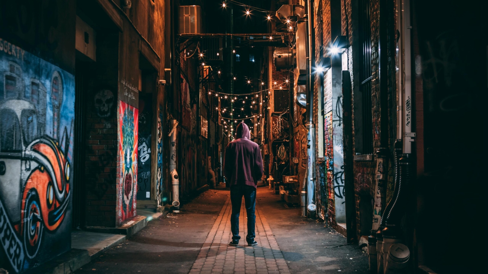 A solitary figure stands in a graffiti-lined alley, illuminated by string lights, at night.